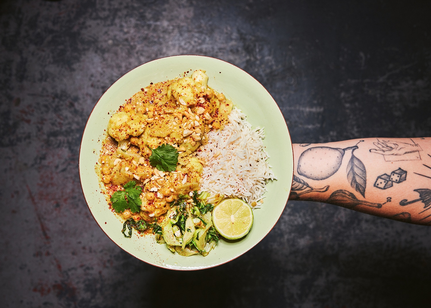 An tattooed arm holding a bowl of whole cauliflower curry
